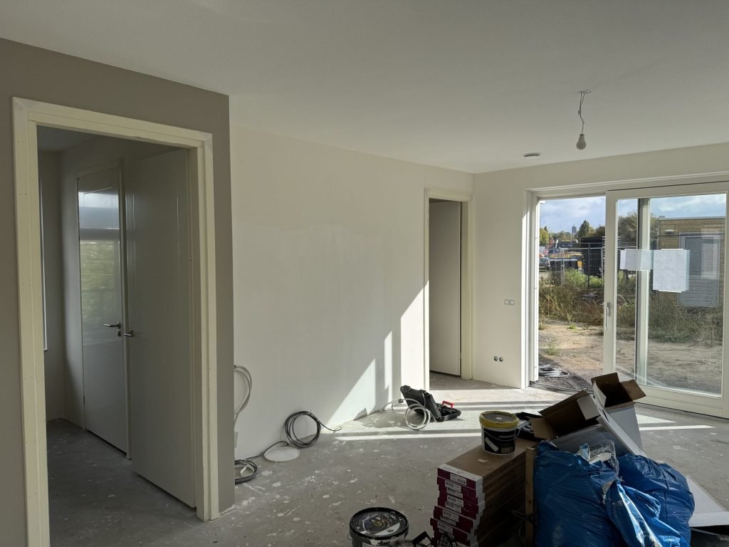 Woonkamer van een nieuwbouwwoning tijdens de afwerking, met verfemmers, kabels en dozen laminaat op de betonvloer