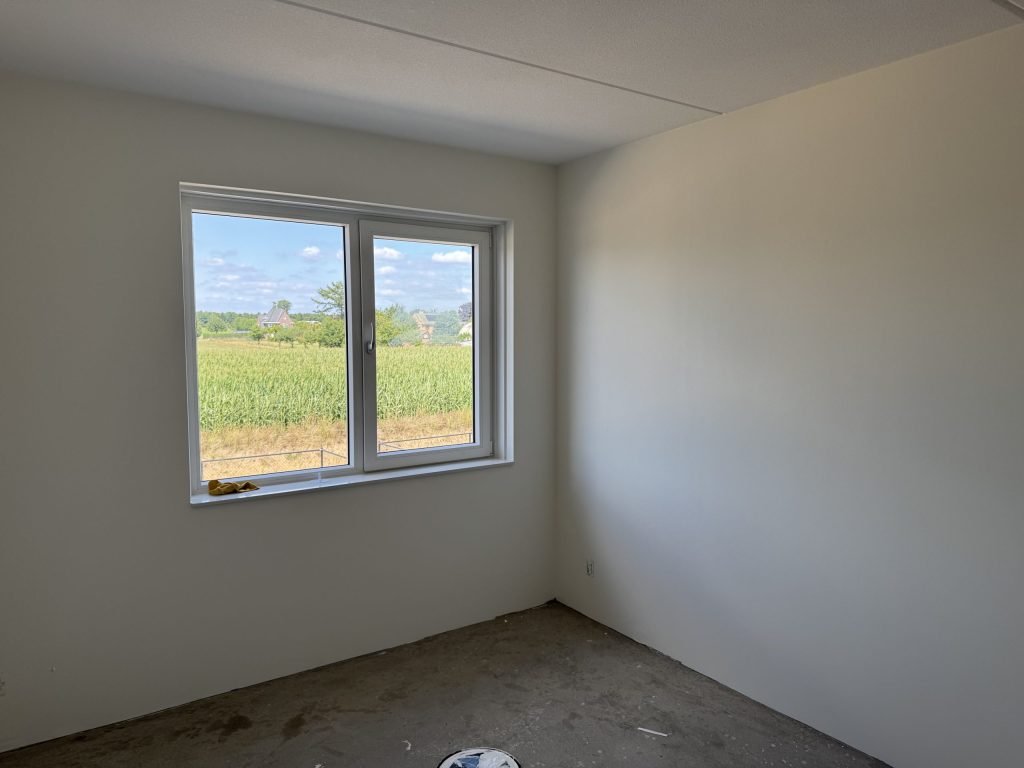 Lege slaapkamer in een nieuwbouwwoning met gladde lichte wanden, een betonvloer en uitzicht op een maisveld